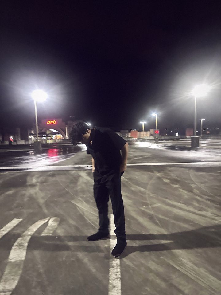 YNGM0M0 dressed in all black standing in an empty parking lot at night under bright lights